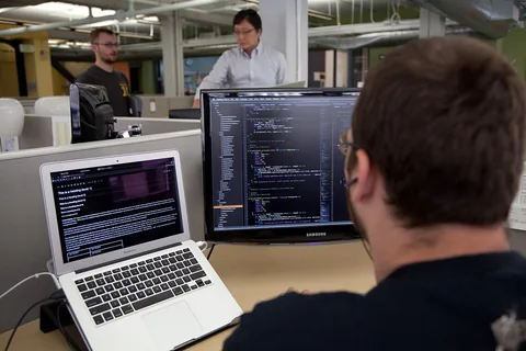 Trevor Parscal coding in the Wikimedia Foundation offices. The image shows the back of a bearded brunette caucasian male with glasses wearing earphones in front of two screens and keyboards