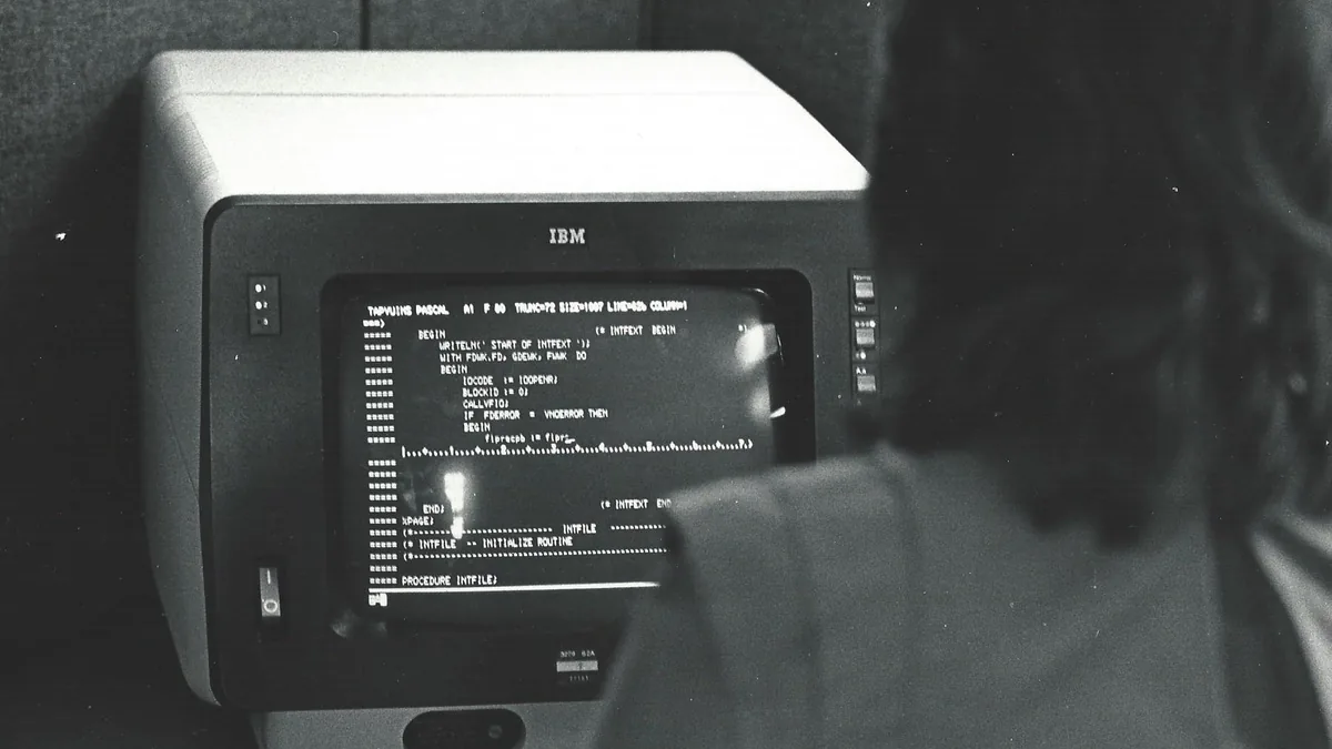An Informatics General computer programmer working in a cubicle at the company's New York City office. She is looking at Pascal language code for the TAPS product on an IBM 3270 terminal